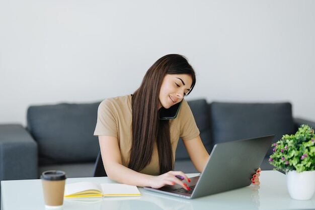 mujer-joven-que-trabaja-computadora-portatil-sonriendo-hablando-telefono-sol-entrando-ventana_231208-13957